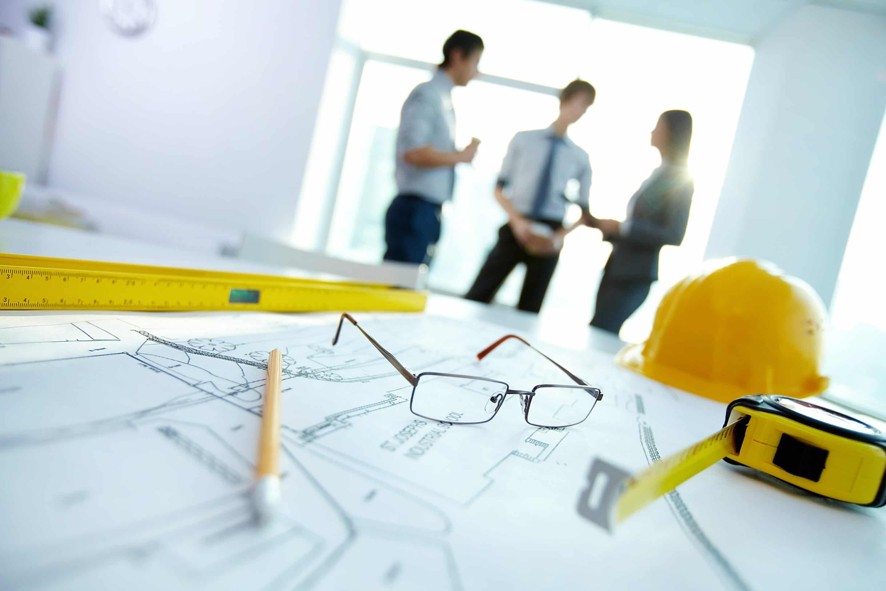 Close up of construction planning papers with three people having a meeting in the background.