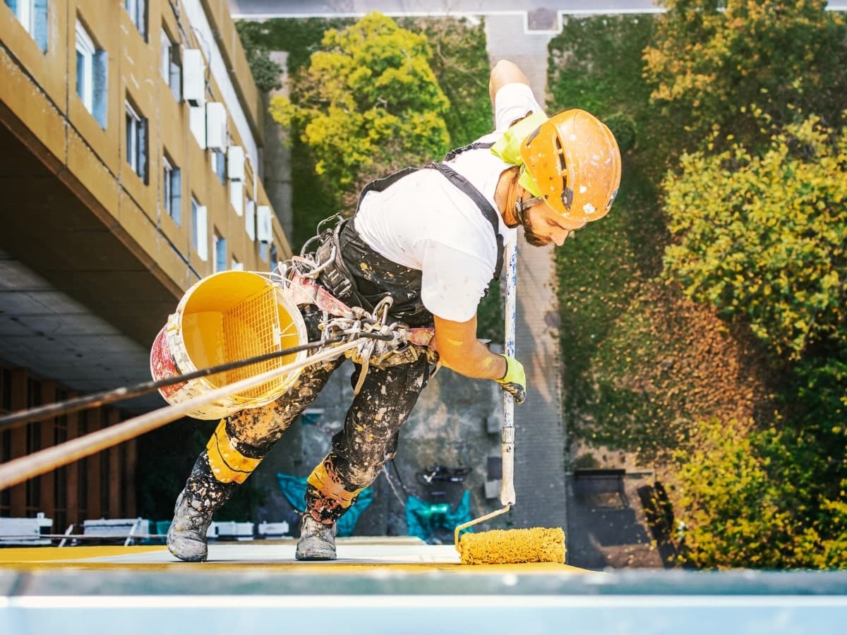 A rope access technician demonstrates working safely at heights, secured with harness and helmet, while applying paint on a building's exterior.