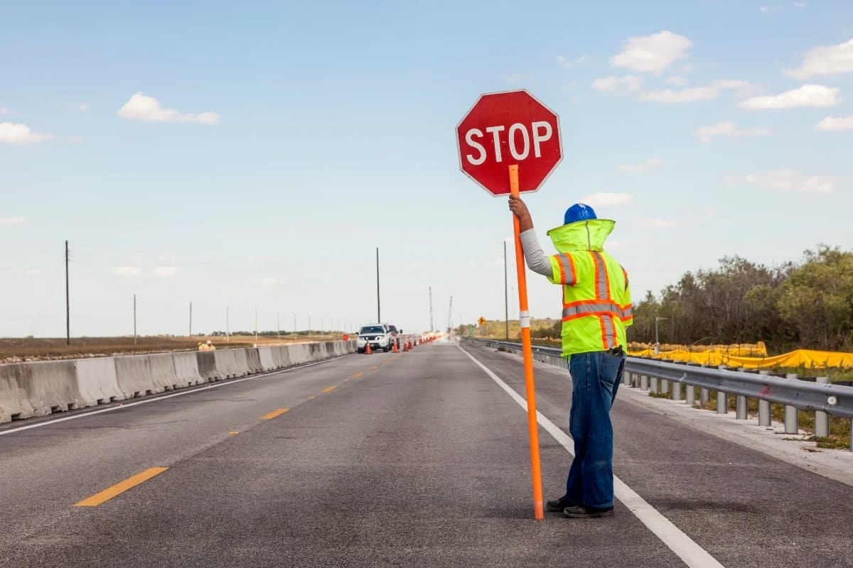 A road worker holding a stop sign, controlling traffic flow with concrete barriers on the side of the road.