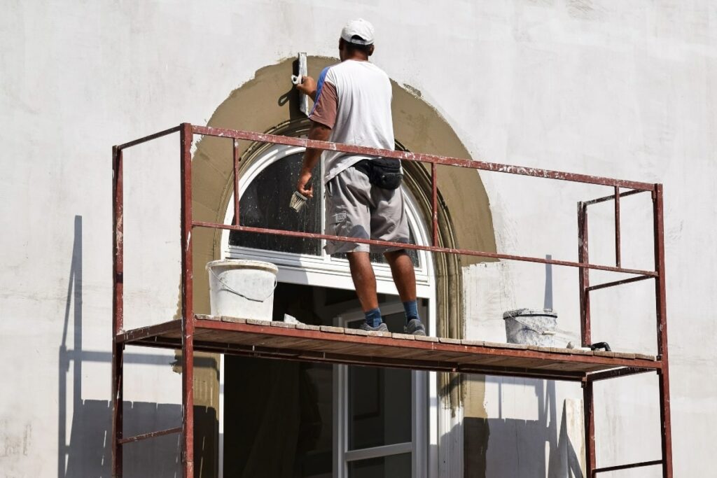 A worker paints a wall while standing on temporary scaffolding outside a building.