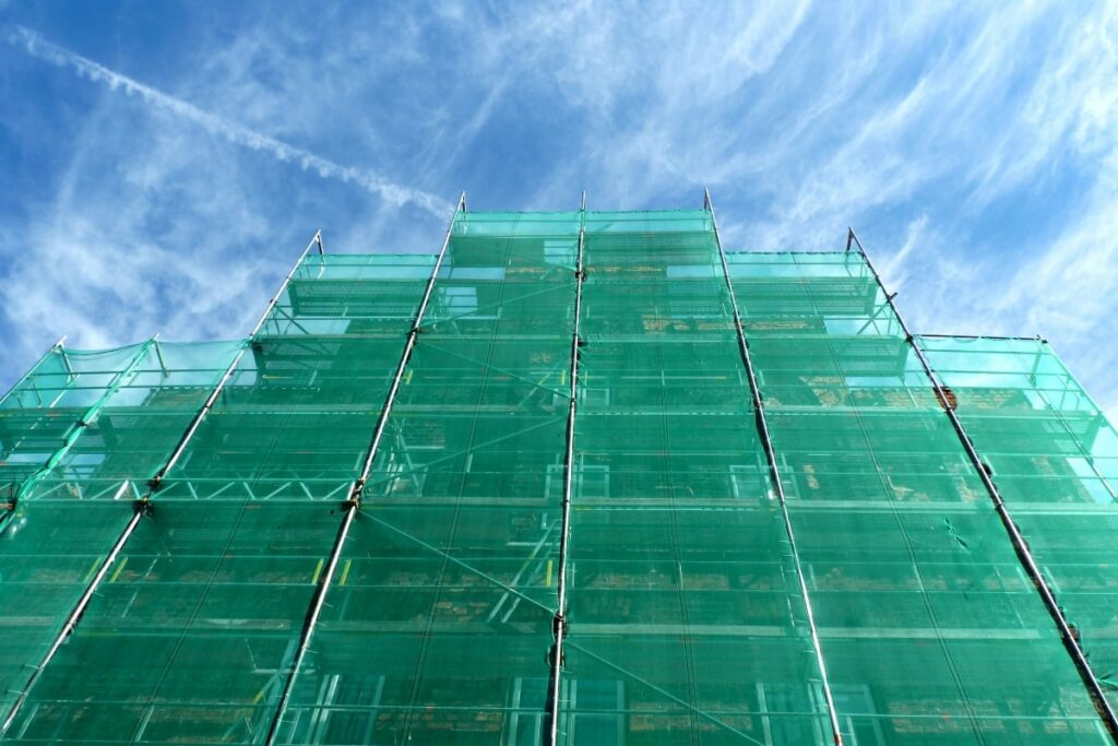 Tall steel scaffolding covered with green safety netting against a blue sky, illustrating its use in large-scale construction projects for enhanced worker safety.