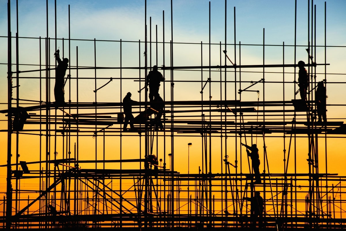 Multiple construction workers silhouette against a sunset, busily working on a complex structure, illustrating various scaffolding types used in building projects.