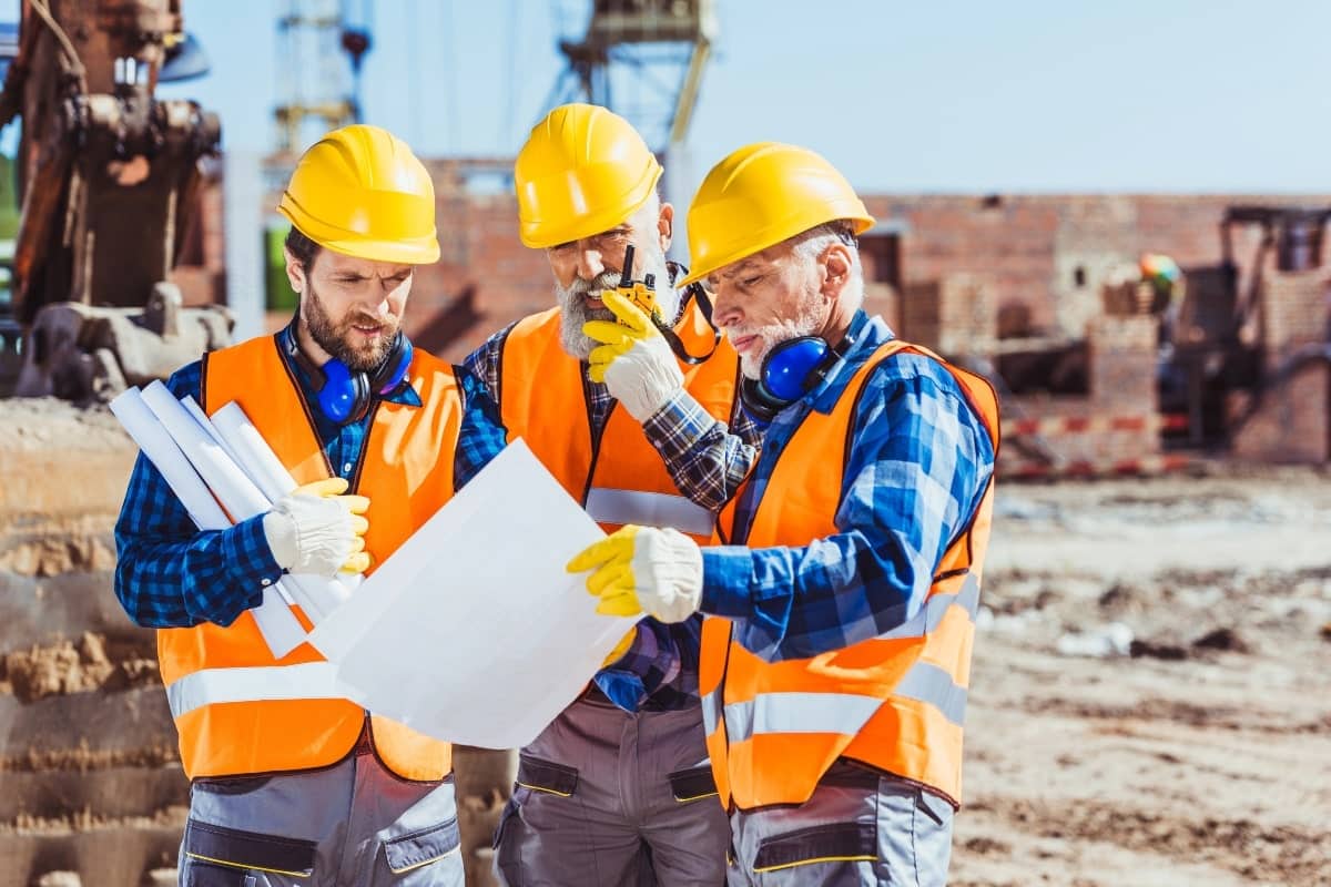 Three men in yellow hardhats, looking a building specifications on a construction site.