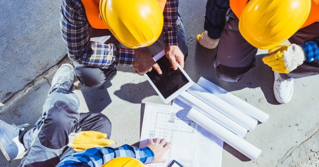 Workers in yellow hard hats gathered around construction plans.